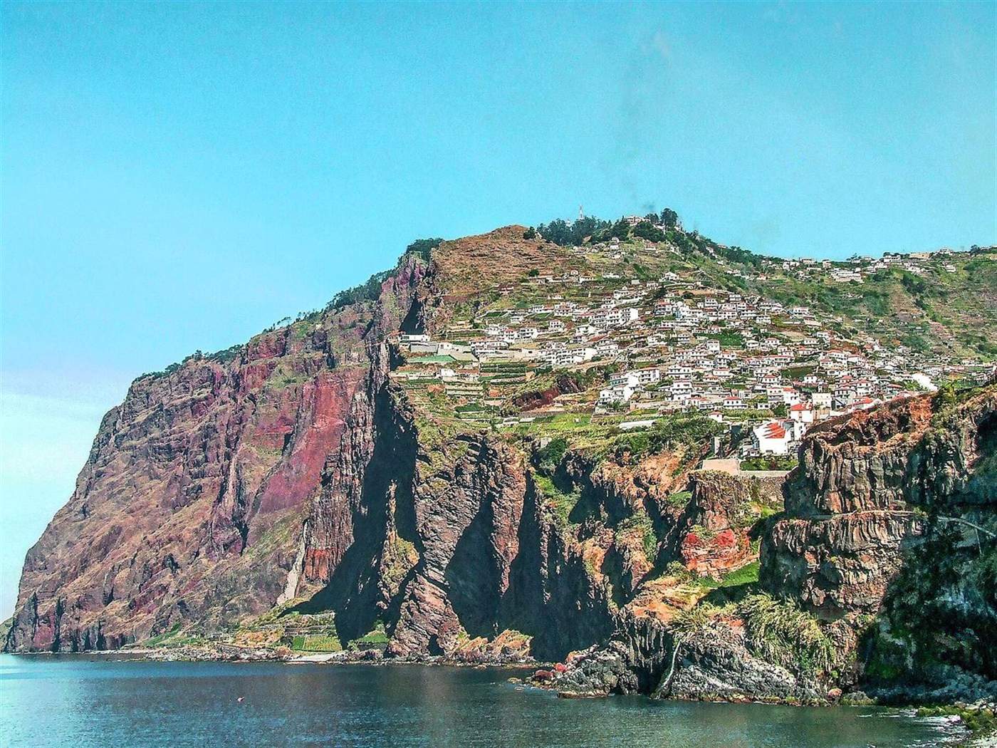 Steile roodbruine kliffen van Cabo Girão met een dorp op de heuvel, gezien vanaf de zee bij Câmara de Lobos