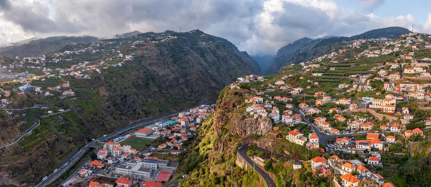 Aerial view of Ribeira Brava built on the slopes at the mouth of a deep valley, Madeira, with a road winding around a rocky outcrop