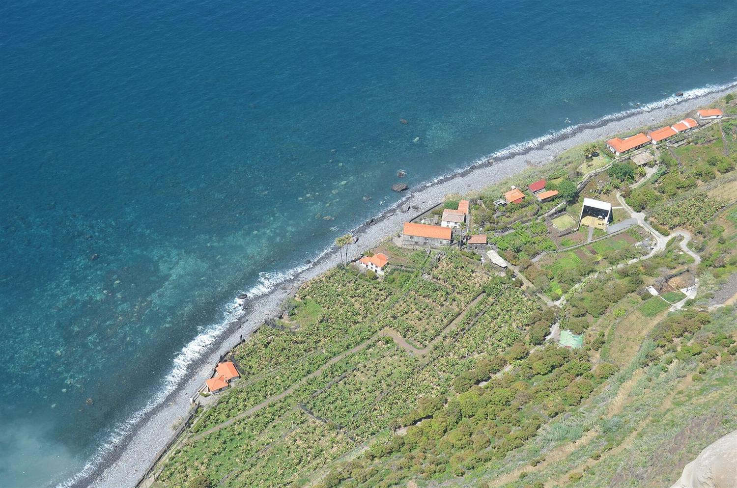 Luftaufnahme einer terrassierten Küstenlandschaft mit Häusern mit orangefarbenen Dächern entlang eines Kiesstrands auf Madeira