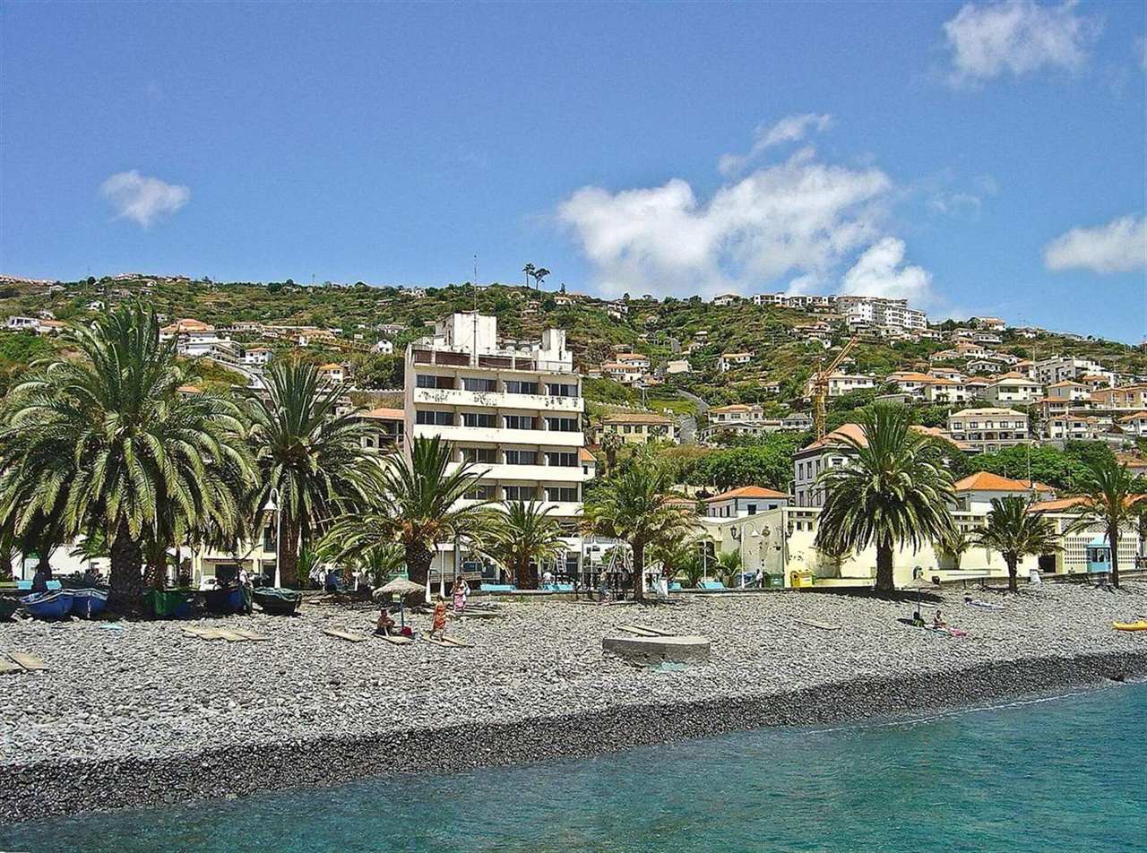 La plage de galets et le front de mer bordé de palmiers à Santa Cruz, Madère, avec des bateaux de pêche tirés sur le rivage et la ville qui s'élève sur la colline derrière