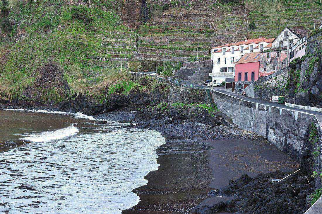 Zwart vulkanisch zandstrand bij Seixal, met terrasvormige wijngaarden op de klif erachter en huizen langs de kustweg, Madeira