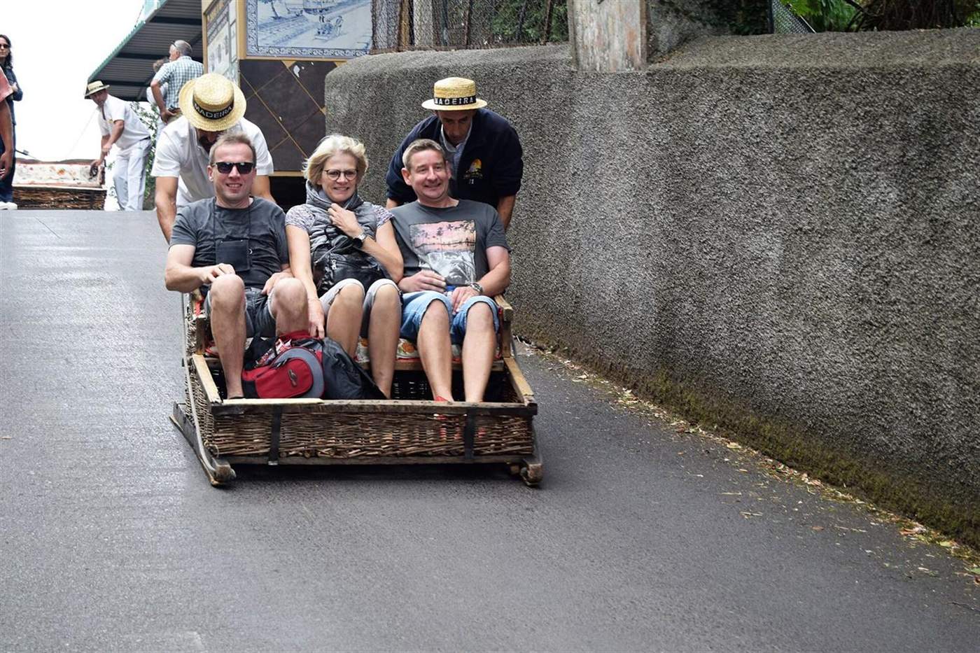 Rieten slee met drie lachende passagiers, geduwd door twee met hoed geklede bestuurders in Monte, Funchal, Madeira