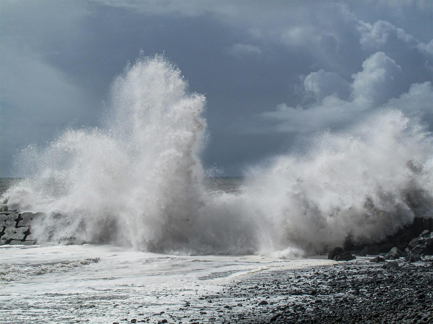 Massive Welle bricht über einem felsigen Wellenbrecher vor stürmischem Himmel auf Madeira, Portugal